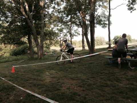 A person riding a bicycle on a grassy area surrounded by trees, with picnic tables in the background. Some caution tape is set up on the ground, and another person is seated at a table nearby. The scene is outdoors, illuminated by sunlight. England Idlewild Mountain Biking Park mountain bike trail.