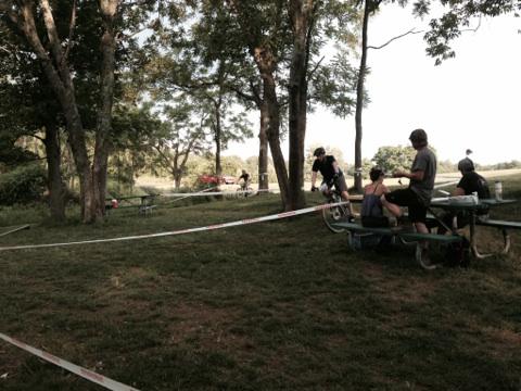 A park scene featuring a grassy area with several trees. People are gathered around picnic tables, while others are riding bicycles and navigating a course marked by tape. The setting suggests an outdoor event or gathering in a casual, natural environment. England Idlewild Mountain Biking Park mountain bike trail.