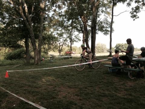 A cyclist rides through a park area, marked by cones and tape, while a group of people sits at picnic tables in the background. Trees provide shade in the outdoor setting, suggesting a recreational event. England Idlewild Mountain Biking Park mountain bike trail.
