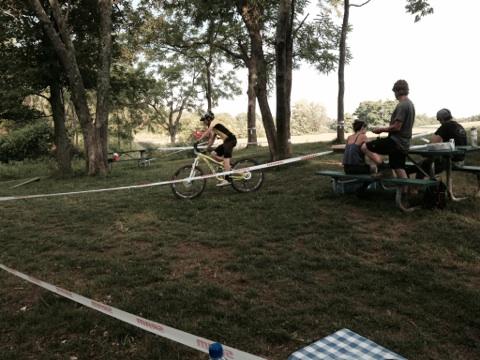 A cyclist in a yellow shirt rides a mountain bike around a grassy area marked with caution tape. In the background, several people are seated at picnic tables under trees, enjoying the outdoors on a sunny day. England Idlewild Mountain Biking Park mountain bike trail.