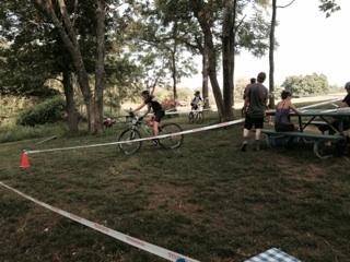A cyclist navigates a course marked by tape in a park setting, while spectators watch from a nearby picnic table. Trees and a grassy area are visible in the background. England Idlewild Mountain Biking Park mountain bike trail.