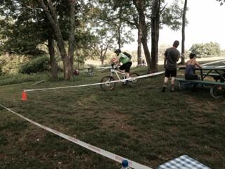 A cyclist wearing a green jersey rides a bicycle along a marked course with cones, while spectators and participants relax near picnic tables in a grassy area surrounded by trees. England Idlewild Mountain Biking Park mountain bike trail.