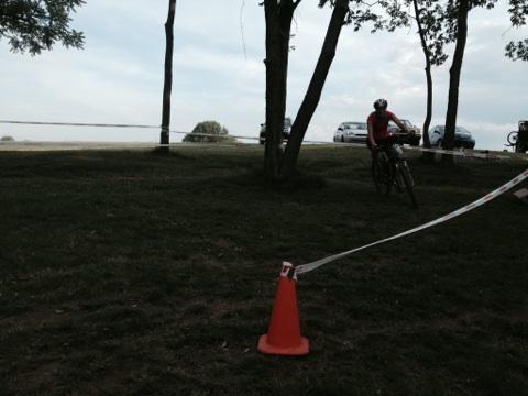 A mountain biker in a red shirt rides on a grassy path near trees, with a traffic cone in the foreground and parked cars in the background. A course marker ribbon is visible, indicating a biking event. The sky is overcast. England Idlewild Mountain Biking Park mountain bike trail.
