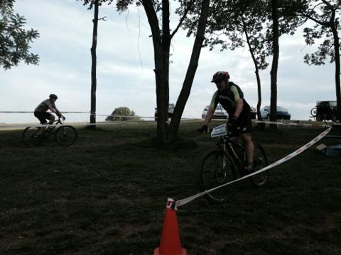 Two cyclists riding mountain bikes on a grassy area, with trees in the background. One cyclist is in the foreground, wearing a helmet and a cycling outfit, while another cyclist can be seen in the background. A traffic cone marks part of the biking trail, and a string barrier is visible. The scene is set in an outdoor environment with an overcast sky. England Idlewild Mountain Biking Park mountain bike trail.