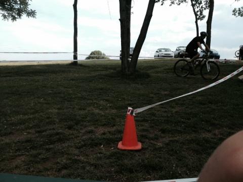A cyclist riding past a traffic cone on a grassy area, with trees in the background and cars parked in the distance. The sky is cloudy, and there is a sense of an outdoor event or race taking place. England Idlewild Mountain Biking Park mountain bike trail.