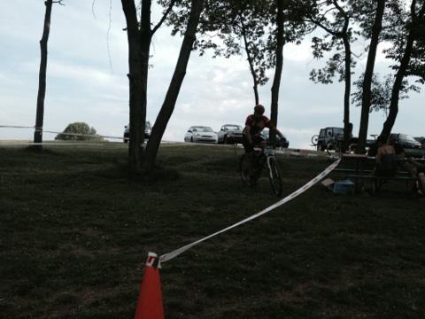 A cyclist rides through a grassy area lined with trees, while a race tape marks the course. In the background, parked vehicles are visible along a path, and a few spectators are seated at a picnic table. The sky is partly cloudy, suggesting an outdoor event. England Idlewild Mountain Biking Park mountain bike trail.