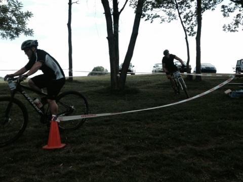 Two mountain bikers navigate a course marked with tape and cones on a grassy area near trees. One cyclist is riding past a cone, while the other is in the background, approaching another turn. Vehicles are visible in the distance, suggesting an outdoor event or competition. England Idlewild Mountain Biking Park mountain bike trail.