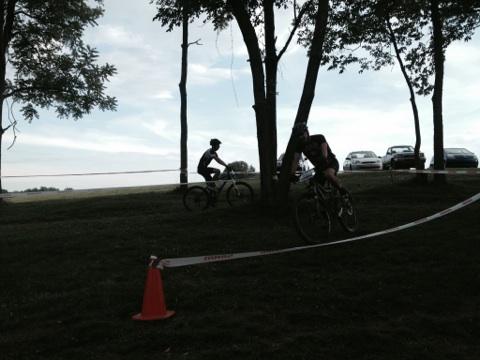 Two cyclists ride along a grassy path surrounded by trees, with a line of cars parked in the background. The scene is set against a cloudy sky, and a traffic cone is visible in the foreground, marking the route. England Idlewild Mountain Biking Park mountain bike trail.