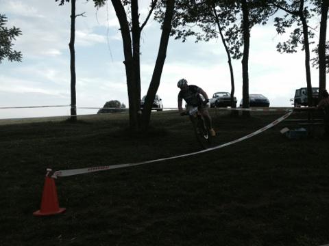 A cyclist navigates a turn on a grassy track marked with tape and cones, surrounded by trees and parked cars in the background under a cloudy sky. England Idlewild Mountain Biking Park mountain bike trail.