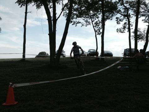 A cyclist navigating a course marked by tape, with trees and parked cars in the background. The scene is set in an outdoor area with a cloudy sky. England Idlewild Mountain Biking Park mountain bike trail.