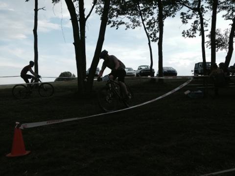 Two cyclists are racing along a grassy path, with trees on one side and parked cars in the background. One cyclist, wearing a helmet and riding a mountain bike, is maneuvering around a course marked by tape and a traffic cone. The scene is set under a cloudy sky, creating a slightly silhouetted effect. England Idlewild Mountain Biking Park mountain bike trail.