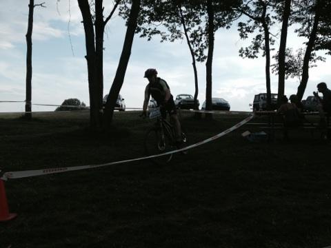 Silhouette of a cyclist navigating a course marked by tape, surrounded by trees. In the background, several parked cars and people sitting at picnic tables are visible under a cloudy sky. England Idlewild Mountain Biking Park mountain bike trail.