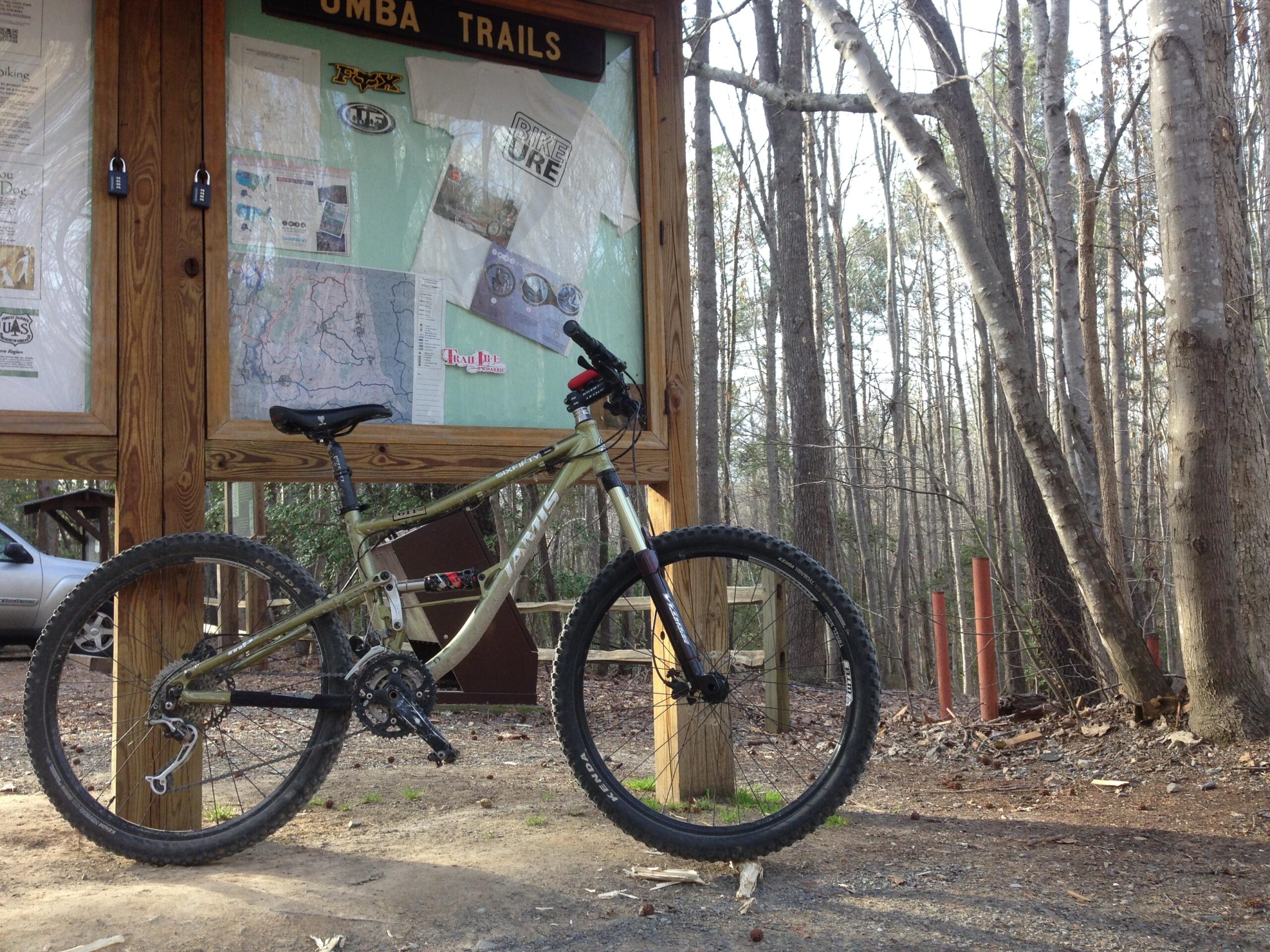 Jamis Dakar Sixfifty B1: A mountain bike is parked next to an information board at a trailhead surrounded by trees. The board features maps and documents related to mountain biking and local trails. A car is visible in the background, and the setting is a wooded area, suggesting a popular outdoor recreation spot.