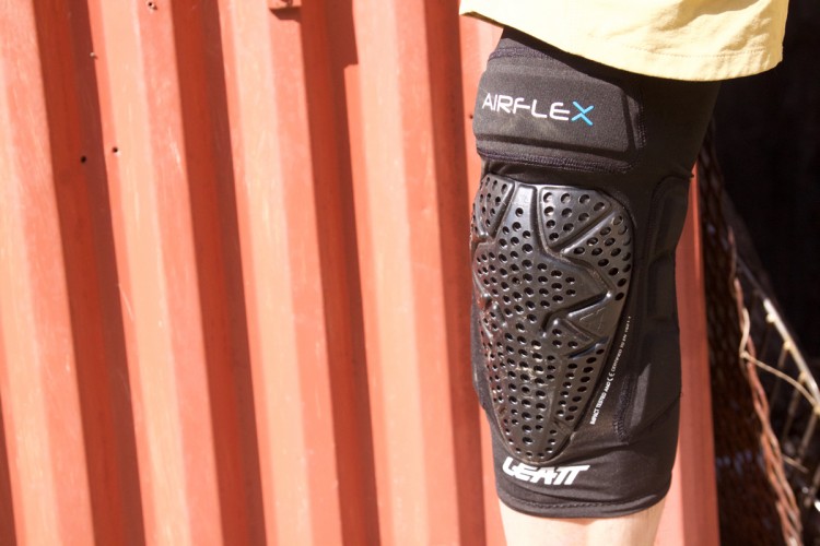 A close-up view of a knee guard featuring a flexible, padded design with ventilation holes, worn on a person's leg. The garment is black with blue and white branding, set against a red corrugated background.