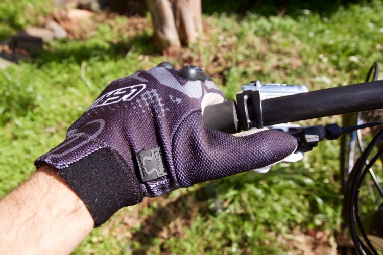 A close-up of a hand wearing a black and gray patterned biking glove, gripping a bicycle handlebar. The background features green grass and blurred foliage.