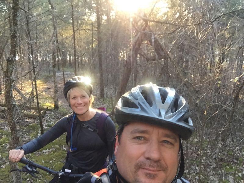 A smiling couple wearing helmets poses for a selfie while mountain biking on a wooded trail. The sun shines through the trees in the background, creating a warm, sunny atmosphere. Jones Mill mountain bike trail.