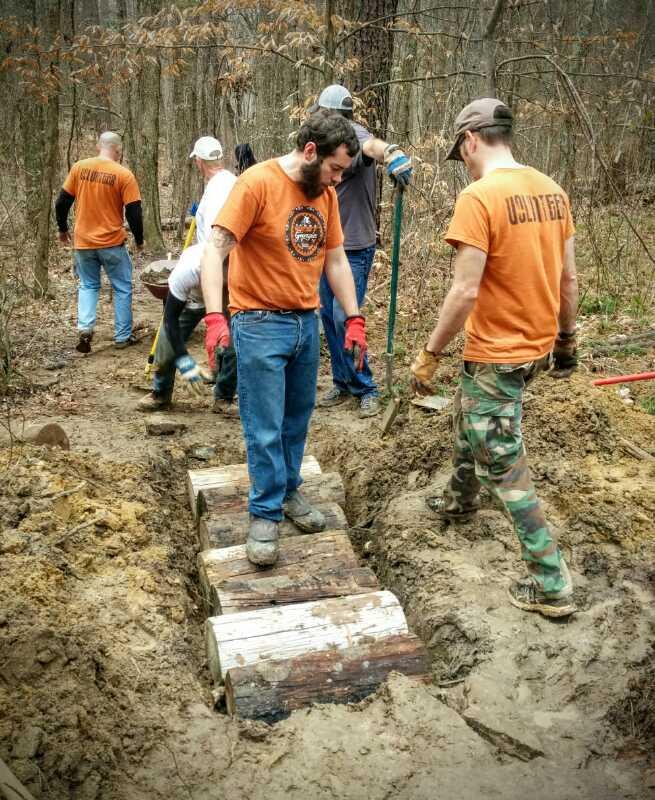 A group of volunteers working together on a dirt trail project in a wooded area. Some individuals are using tools like shovels and picks, while others are positioning logs to create a walkway. Everyone is wearing gloves and casual clothing, with several attendees in matching orange shirts. The environment is natural with bare trees and soil visible around the work site. Clinton Nature Preserve mountain bike trail.