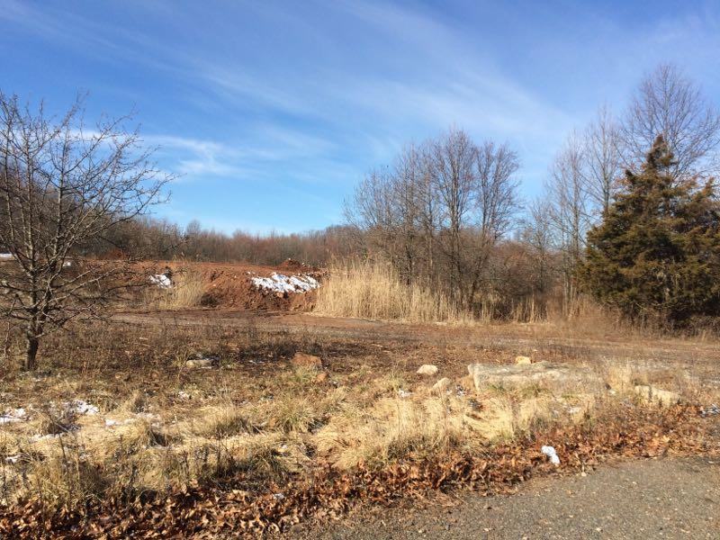 A scenic view of a rural landscape featuring a mix of dry grass, scattered rocks, and sparse trees. In the background, a mound of dirt is visible, and patches of snow are scattered across the ground. The sky is bright blue with a few wispy clouds. Long Pond mountain bike trail.