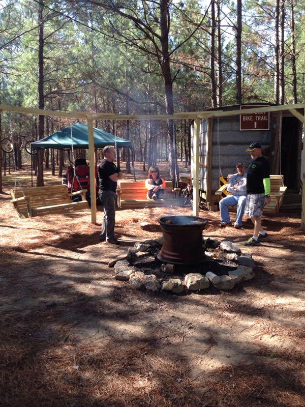 A group of people gathered around a fire pit in a wooded area, with smoke rising from the pit. There are wooden swings and benches nearby, and the scene is surrounded by tall pine trees. Some individuals are standing while others are seated, enjoying the outdoors. A sign indicating a bike trail is visible in the background. Mt. Zion Bike Trails mountain bike trail.