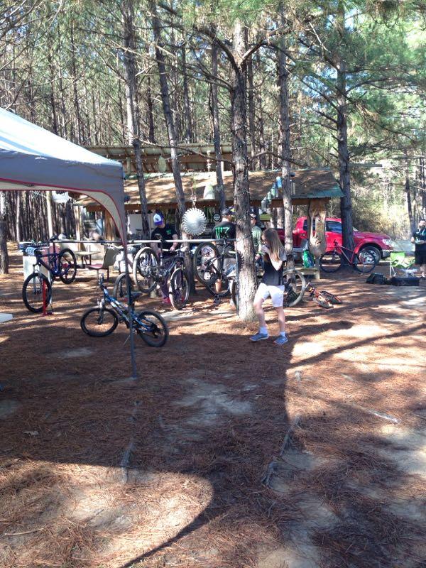 A wooded outdoor area with tall pine trees, featuring a gathering space with a tent. Several bicycles are parked nearby, and a person is seen walking next to the trees. In the background, there is a building with a wooden structure and a red truck parked. Sunlight filters through the trees, casting shadows on the ground. Mt. Zion Bike Trails mountain bike trail.
