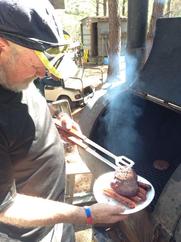 A person wearing a black shirt and a cap is grilling food outdoors. They are using tongs to lift a burger pattie from a smoky barbecue grill while holding a plate with sausages. Trees and a shed are visible in the background, suggesting a camping or outdoor setting. Mt. Zion Bike Trails mountain bike trail.