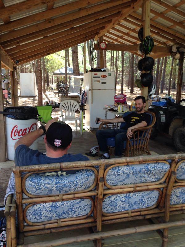 Two men are seated in a rustic outdoor space with a roof supported by wooden beams. One man, seen from behind, is wearing a dark cap and a gray shirt. The other man sits comfortably in a wicker chair, wearing a black shirt and jeans, smiling as they engage in conversation. In the background, there's a small kitchenette area with a refrigerator, trash can, and various items hanging from hooks. The setting is surrounded by tall trees, indicating a natural, relaxed atmosphere. Mt. Zion Bike Trails mountain bike trail.