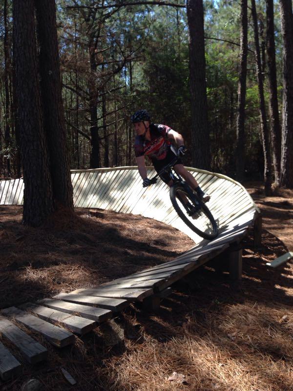 A mountain biker riding on a wooden track through a forest, navigating a curved section of the path surrounded by trees and pine needles. The cyclist is wearing a helmet and a brightly colored jersey. Mt. Zion Bike Trails mountain bike trail.
