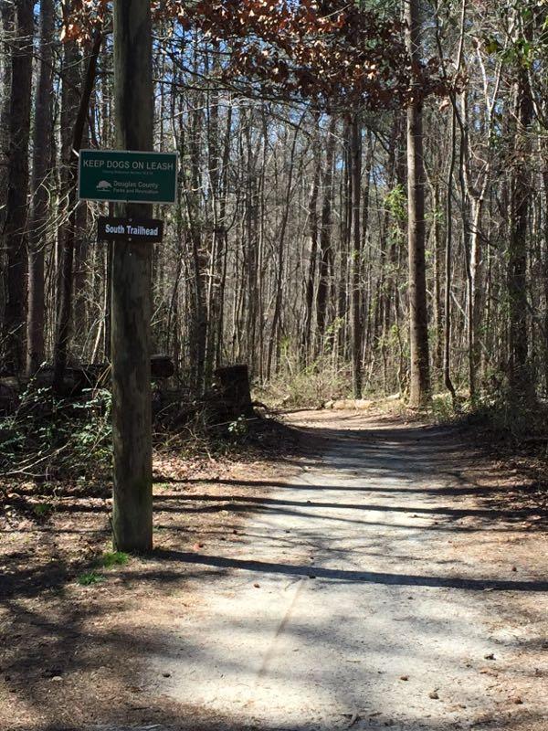 A dirt path winding through a wooded area, marked by a sign that instructs to keep dogs on a leash and indicates the South Trailhead. The scene is sunlit, with trees lining either side of the path. Clinton Nature Preserve mountain bike trail.