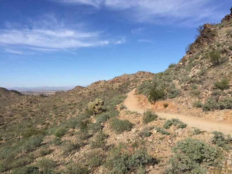 A winding dirt path trails through rocky terrain, surrounded by sparse vegetation and rolling hills under a clear blue sky. The landscape features a mix of shrubs and desert plants, creating a natural environment typical of arid regions. San Tan mountain bike trail.