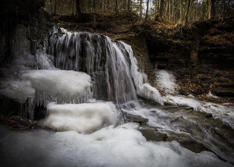 A frozen waterfall cascading over rocky ledges, surrounded by icicles and patches of snow, with a backdrop of trees and natural foliage. The scene captures the serene beauty of winter in a forest setting. Thatcher State Park mountain bike trail.