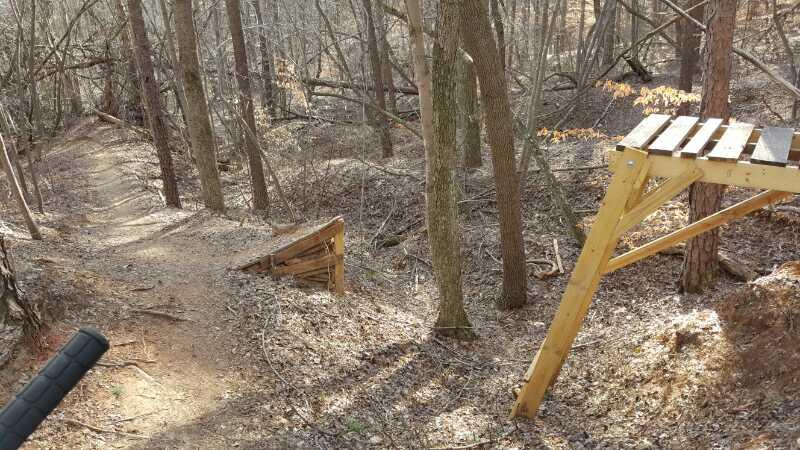 A dirt bike trail winding through a wooded area, featuring a wooden bike ramp on the right side and a small jump constructed with wooden planks on a slope. The surrounding environment is mostly bare trees, with some foliage on the ground, suggesting early spring or late fall conditions. Salem Lake mountain bike trail.