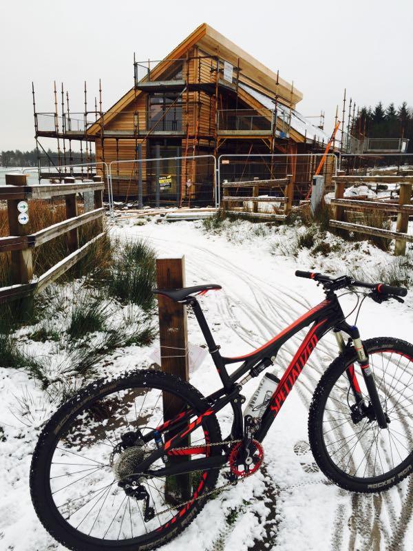 A mountain bike leaning against a wooden post in a snowy landscape, with a partially constructed wooden house in the background surrounded by scaffolding and fencing. Falkirk xc mountain bike trail.