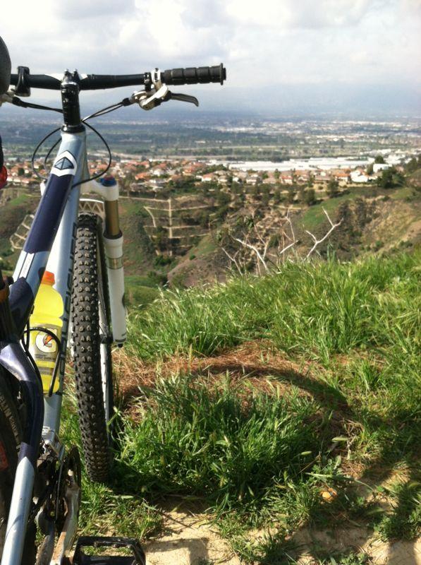 Mountain bike parked on grass overlooking a scenic valley with houses and distant hills under a partly cloudy sky. Turnbull Canyon mountain bike trail.