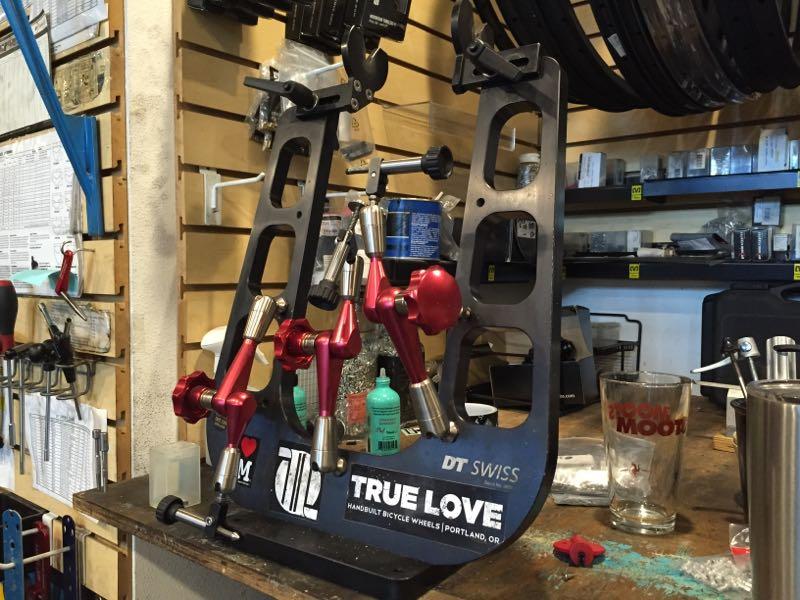 A bicycle wheel truing stand with various tools and components visible, including red and silver adjustment arms. The stand is placed on a workbench surrounded by bottles of bike maintenance supplies and a glass. Behind it is a wooden wall with organized tools and bike parts neatly displayed.