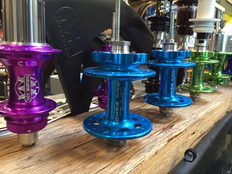 A close-up photograph of several colorful bicycle hubs displayed on a wooden surface. The hubs are in various shades, including purple, blue, and green, with metallic finishes. They are part of a bike shop display, showcasing different designs and brands.
