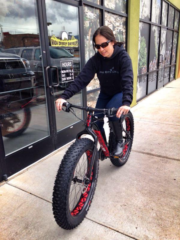 A person wearing sunglasses and a black hoodie is riding a fat tire bike outside a bike shop. The bike has thick, knobby tires and a black and red frame. The shop's glass door displays the store hours. The setting appears to be a sidewalk with a cloudy sky in the background.