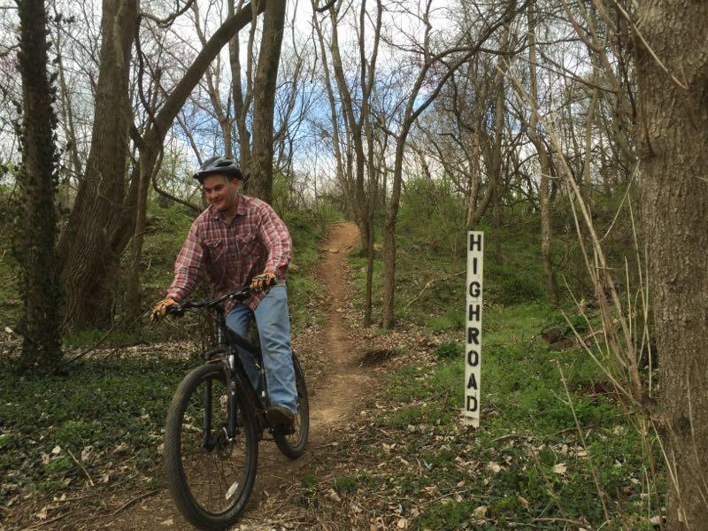 A person riding a mountain bike on a dirt trail surrounded by trees, with a sign labeled "HIGHROAD" nearby. The cyclist is wearing a helmet and a plaid shirt, smiling as they navigate the path. The scene is set in a lush, green forest with a partly cloudy sky. Low Hollow mountain bike trail.