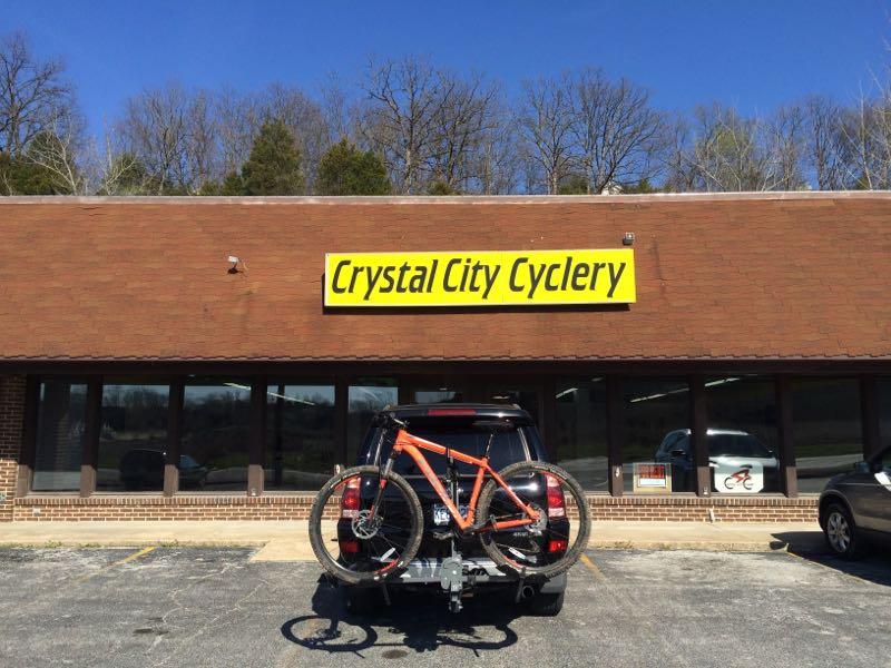Front view of the Crystal City Cyclery shop, featuring a large yellow sign. An orange mountain bike is mounted on the back of an SUV parked in front of the store. The building has large windows, and it