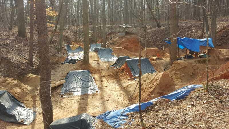 A serene forest scene featuring several covered excavation sites in a wooded area. Each site is partially covered with blue tarps and surrounded by trees, with exposed sandy soil and some shaded patches. The sunlight filters through the branches, creating a peaceful atmosphere in the natural setting. Salem Lake mountain bike trail.