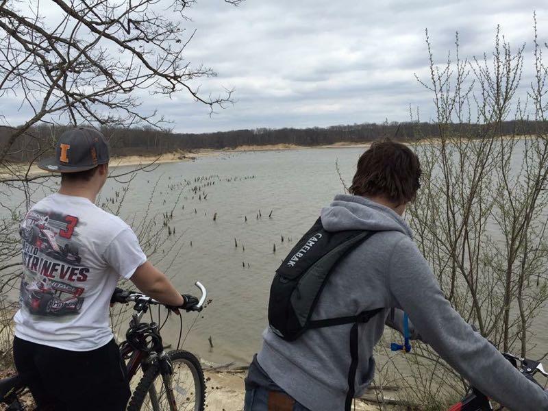 Two young men are standing on a rocky ledge overlooking a body of water. They are positioned beside bicycles. The sky is overcast, and there are trees in the foreground. In the water, there are remnants of submerged trees or stumps visible. The landscape in the background features a shoreline with sparse vegetation. Camp Camfield mountain bike trail.