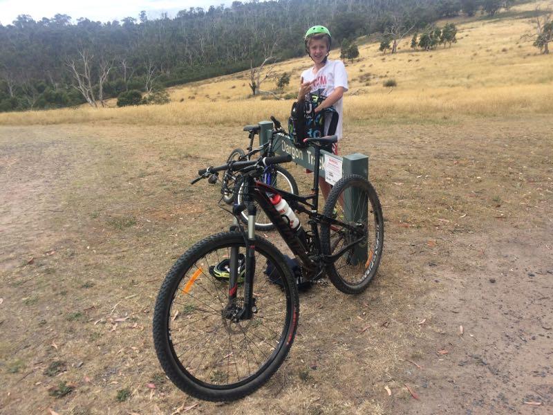 A young boy wearing a helmet and casual clothing stands next to a mountain bike, smiling and giving a thumbs-up. The bike is positioned near a trail sign labeled "Jargon Trail," with a grassy field and trees in the background. The scene captures a moment of outdoor adventure and enthusiasm for biking. Lysterfield Mountain Bike Area mountain bike trail.