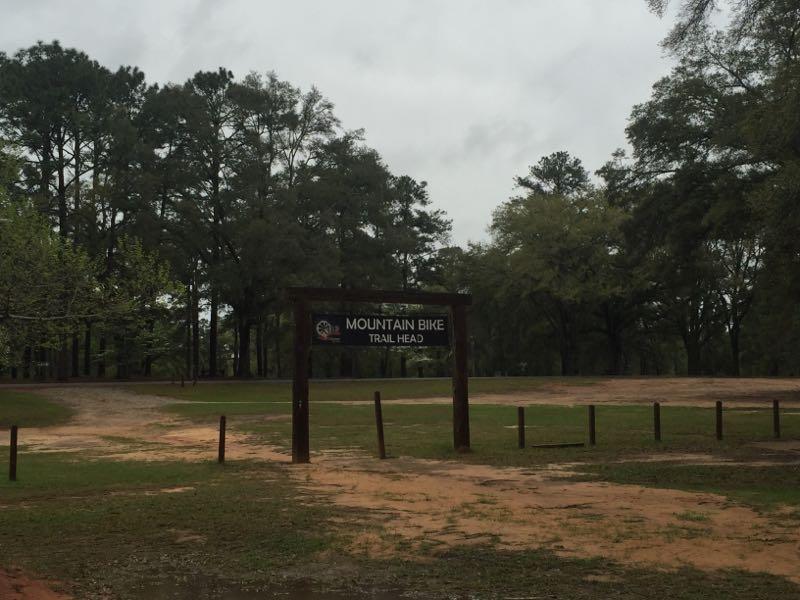 Sign for a mountain bike trail head, set in a wooded area with a cloudy sky. The sign is displayed prominently in the foreground, and the surrounding landscape features patches of grass and dirt paths leading into the trees. Chehaw Park mountain bike trail.