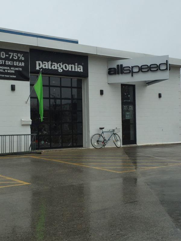 Exterior view of a retail store featuring signage for "Patagonia" and "allspeed." A green flag is displayed in front, and a bicycle is secured to a railing on the sidewalk. The scene is set on a rainy day, indicated by wet pavement and overcast skies.