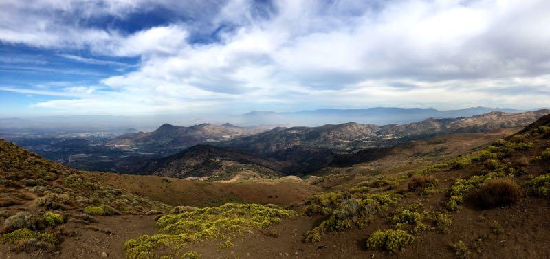 A panoramic view of a mountainous landscape under a partly cloudy sky. The foreground features rugged terrain with patches of green vegetation, while the background reveals rolling hills and distant mountains shrouded in a soft haze. The scene captures the beauty of nature, with varying shades of brown and green across the terrain. Paso del Diablo / Tirabuzon mountain bike trail.