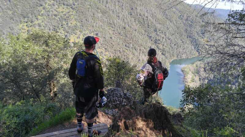 Two individuals standing on a rocky ledge overlooking a scenic river valley, surrounded by lush greenery and rolling hills. The person on the left is wearing a backpack and a baseball cap, while the person on the right has a red backpack and is dressed in outdoor gear. The river winds through the valley below, reflecting the sunlight. Clementine / Forresthill Connector Trail mountain bike trail.