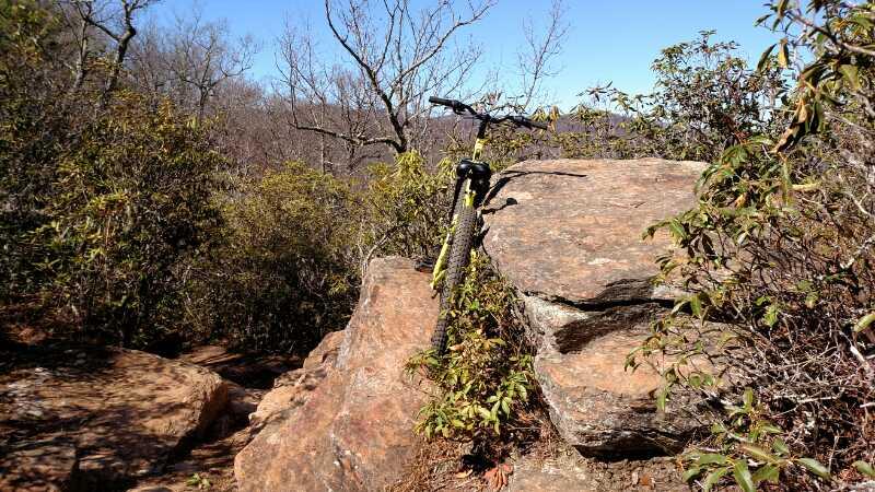 A mountain bike leaning against a large rock surrounded by sparse vegetation and trees under a clear blue sky. Bennett Gap / 138 mountain bike trail.