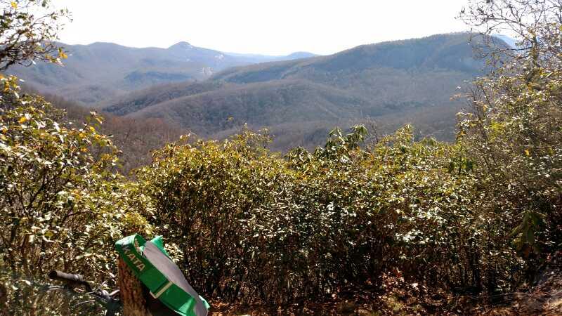 A scenic view of mountains in the background, featuring rolling hills covered in trees and shrubs, with a clear sky above. In the foreground, there is a partially visible bicycle leaning against a bush, adding a sense of adventure to the landscape. Bennett Gap / 138 mountain bike trail.