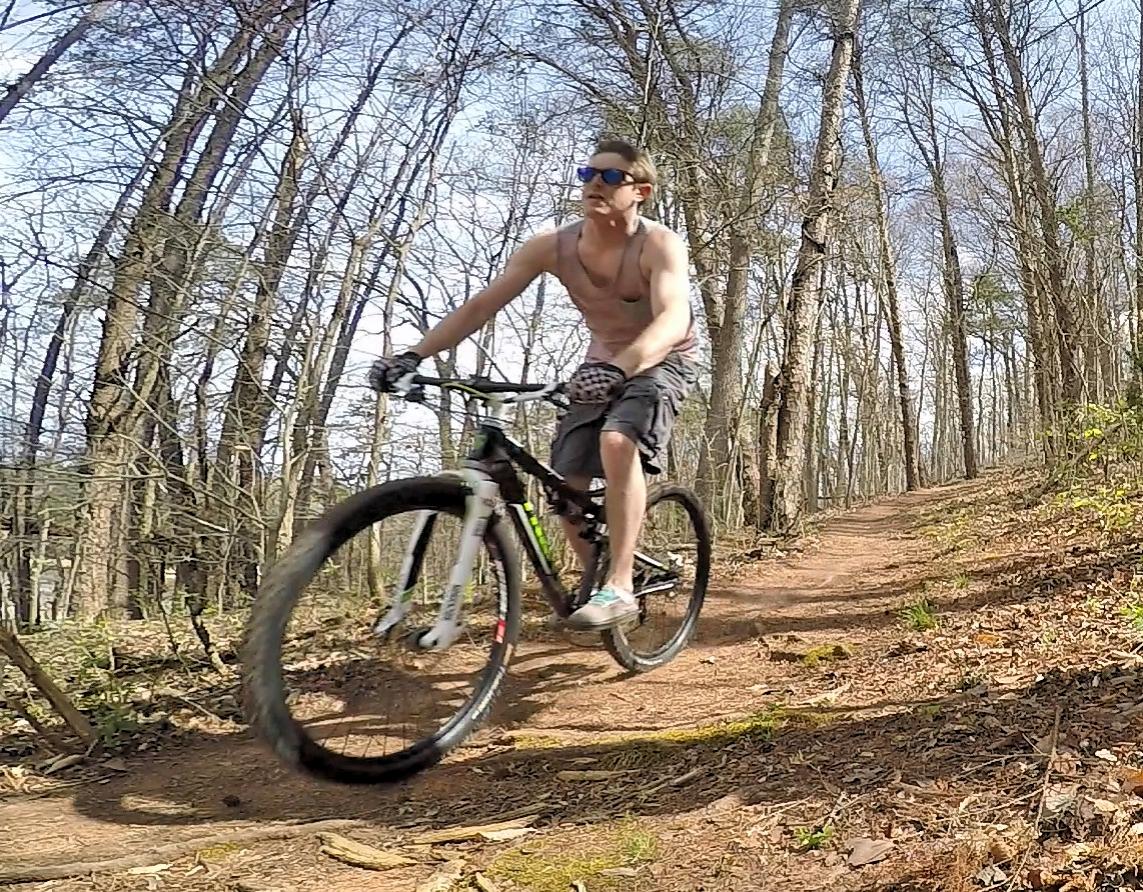 Trek Superfly Full Suspension: A person riding a mountain bike on a dirt trail through a forest, wearing shorts and sunglasses, with trees in the background and a clear blue sky above.