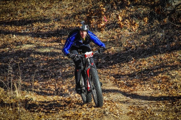 A cyclist in a blue jacket rides a fat bike along a leaf-covered trail, navigating through a wooded area during autumn.