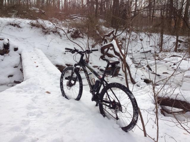A mountain bike parked on a snow-covered path next to a small stream in a wintery forest landscape. The surrounding area is blanketed in snow, with bare trees visible in the background. Trails seperated by streets mountain bike trail.